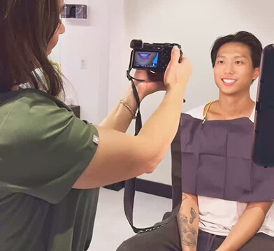 A person in a green shirt takes a photo of a seated individual wearing a dental bib in a modern clinic setting.