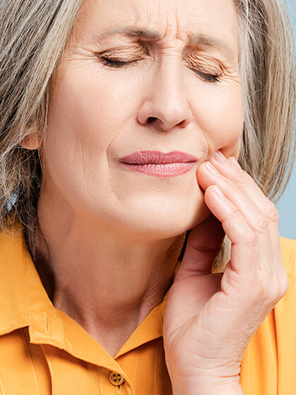 Close-up of an older woman with graying hair wearing a yellow shirt grimacing in pain while holding her cheek, indicating a toothache or jaw discomfort.
